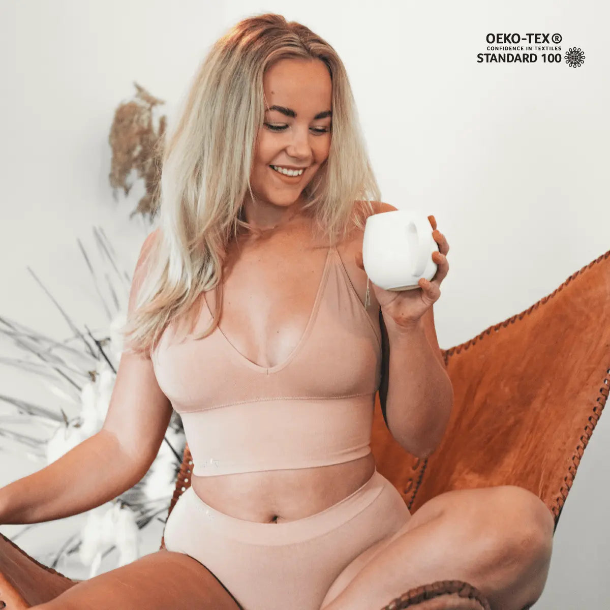 Smiling woman in beige underwear sitting on chair holding white mug promoting self-care for down there.
