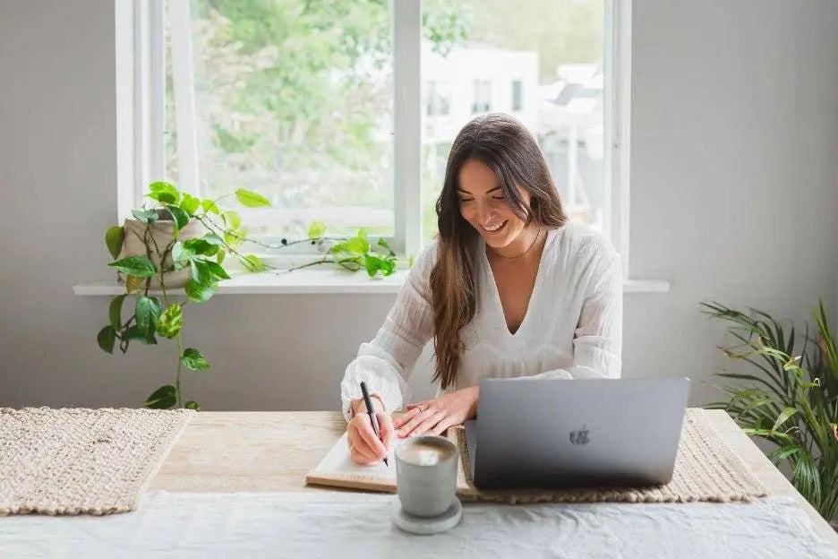 woman writing in journal with laptop and coffee at cozy bright workspace offering winter blues assistance tips