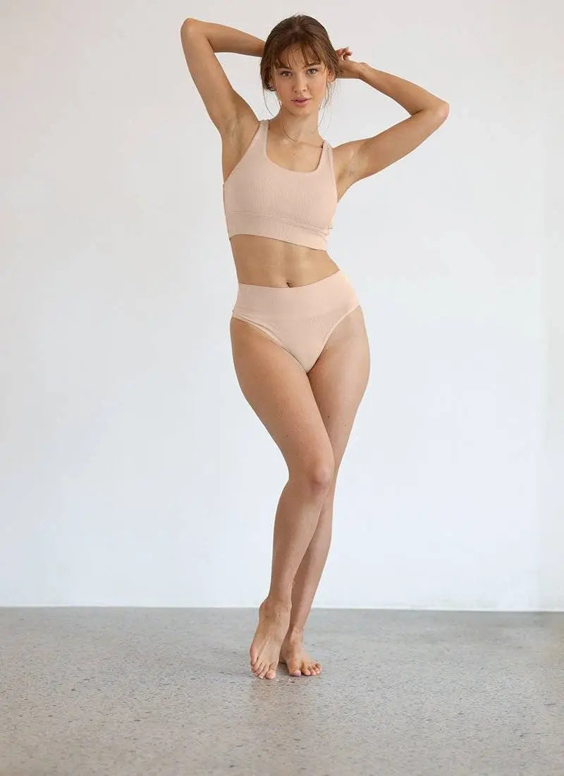 woman wearing nude high-waisted G string and matching crop top standing barefoot against white wall