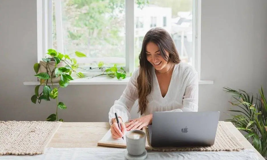 woman writing in journal with laptop and coffee at cozy bright workspace offering winter blues assistance tips