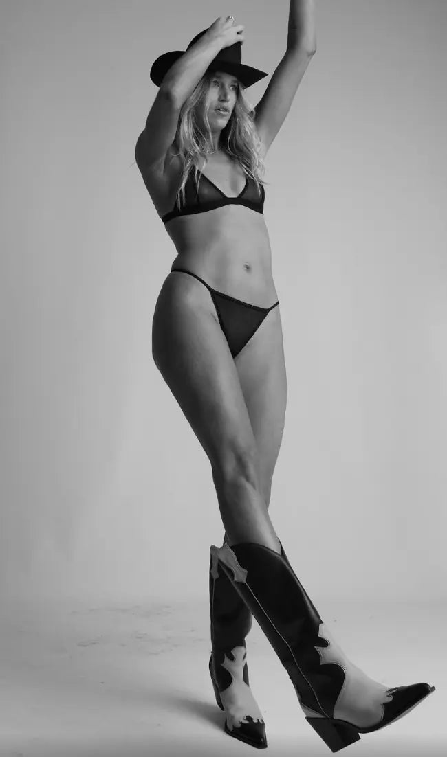 Woman wearing a black G-string, black bra, and cowboy boots with a hat in a studio setting