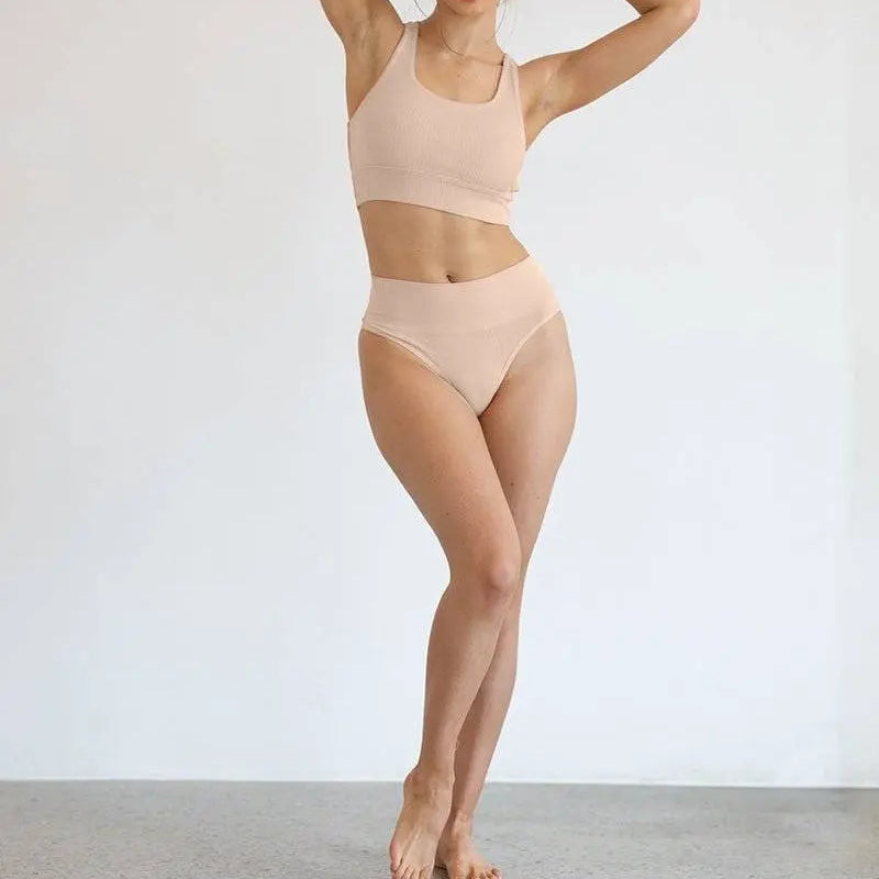 woman wearing nude high-waisted G string and matching crop top standing barefoot against white wall